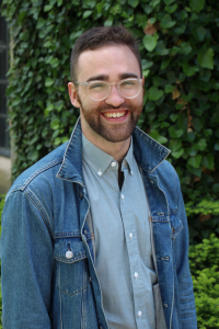 A headshot photo of Michael McGovern who his standing, smiling, and wearing thin framed glasses and a denim jacket over a buttoned shirt