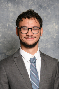 Headshot photo of Colby Thornton, wearing black framed glasses, a gray suit jacket, blue tie, white shirt, and smiling at the camera.